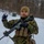 A soldier in digital camouflage uniform is standing in a snowy, wooded environment while holding a rifle. The individual is wearing tactical gear including a plate carrier vest, gloves, and a coyote tan beanie. Snow appears to be falling lightly in the background.
