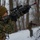 A military service member in MARPAT (Marine digital camouflage) uniform is aiming a scoped rifle upward in a snowy woodland environment. A second service member is visible in the background in a low crouch position, also holding a weapon. The scene appears to depict a winter field training exercise.