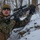 A soldier in woodland digital camouflage (MARPAT pattern consistent with USMC) is positioned on a snow-covered hillside in a winter forest environment. The individual is holding an AR-platform rifle equipped with multiple accessories and appears to be in a tactical training or field exercise scenario. The setting suggests cold-weather operations training.