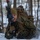 A U.S. Marine in MARPAT woodland camouflage uniform is low-crawling through snow in a wooded winter environment while carrying a rifle and large tactical backpack. The service member is wearing a coyote brown watch cap and tactical gloves, appearing to be engaged in a cold-weather training exercise. The image is a sharp, close-up action shot taken in a snow-covered forest setting.