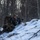 A soldier in camouflage uniform is positioned on a snow-covered hillside in a winter woodland environment, aiming a scoped rifle in a prone/low-profile firing position. The individual is wearing MARPAT (Marine pattern) camouflage and carrying a large tactical backpack. The scene appears to depict a military training exercise or field operation in cold-weather conditions.