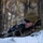 A military or law enforcement personnel is positioned in a prone firing position in a snowy, wooded environment. The individual is partially concealed behind a snow-covered embankment with dry grass, aiming a rifle equipped with optical and tactical accessories. The setting appears to be a cold-weather training or operational scenario.
