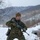 A young soldier in MARPAT (Marine Pattern) digital camouflage uniform is moving through a snow-covered terrain while carrying a rifle. The individual is equipped with full tactical gear including a plate carrier vest, utility belt, and tactical gloves, wearing a coyote brown beanie. The winter environment features bare trees, snow-covered ground, and hilly terrain in the background.
