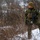 A soldier in digital camouflage uniform moves through a snow-covered wooded area during winter conditions. The individual is equipped with tactical gear including a plate carrier vest and carries a rifle. The scene appears to depict a military field exercise or patrol in a cold-weather environment.