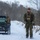 Two military personnel in MARPAT (Marine pattern) camouflage uniforms are operating in a snowy, wooded environment. One service member is walking on a snow-covered trail while carrying a weapon and wearing tactical gear, while a second is seated in a military utility terrain vehicle (UTV). The scene appears to depict a cold-weather training or patrol exercise.