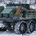 A green military unmanned ground vehicle (UGV) or robotic logistics platform is photographed in a snowy, winter environment. The vehicle features eight large all-terrain tires with aggressive tread patterns and is loaded with military gear including tactical cases, backpacks, and equipment. A WARN winch is visible on the front, indicating recovery capability.