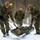 Military personnel in woodland MARPAT camouflage uniforms are conducting a casualty evacuation drill in a snowy, wooded environment. Three soldiers are actively transporting a simulated casualty on a green litter/stretcher across snow-covered ground. The scene appears to be a cold-weather training exercise based on the winter conditions and yellow training markers visible on equipment.