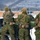 Four military personnel in MARPAT (Marine pattern) camouflage uniforms are handling what appears to be a large munition or rocket near a military vehicle in a snowy winter environment. The soldiers are wearing tan/coyote brown gear, tactical vests, and cold-weather caps. A large orange/tan building and snow-covered mountains are visible in the background.