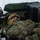 Two military personnel in full combat gear are positioned behind a vehicle in a snowy environment, appearing to be in a tactical prone or low-profile firing position. They are wearing MARPAT (Marine pattern) camouflage uniforms with tactical helmets equipped with night-vision goggle mounts and ballistic eye protection. The scene suggests a cold-weather military training exercise or operational scenario.