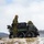 A group of military personnel in digital camouflage uniforms are gathered around a small unmanned ground vehicle (UGV) in a snowy, winter terrain. The vehicle appears to be a tracked robotic platform loaded with equipment cases and gear. The setting suggests a cold-weather military training exercise or field operation.