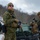 The image shows multiple military personnel in winter combat gear standing near a green armored vehicle in a snowy, wooded environment. The soldiers are wearing digital camouflage uniforms consistent with USMC MARPAT pattern, equipped with tactical vests, backpacks, and rifles. The setting appears to be a cold-weather training exercise or operational deployment.