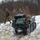 Two military personnel in camouflage uniforms are operating alongside a small unmanned ground vehicle (UGV) on a snow-covered terrain. The tracked robotic vehicle is loaded with military equipment including hard cases and a backpack, navigating a snowy hillside. The setting appears to be a winter training exercise in a wooded area.