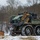 A military service member in MARPAT camouflage uniform stands beside a small unmanned ground vehicle (UGV) in a winter training environment. The vehicle, consistent with a MUTT (Multi-Utility Tactical Transport) or similar robotic logistics platform, is loaded with tactical gear and equipment cases. The setting appears to be a military training area with light snow cover and bare deciduous trees in the background.