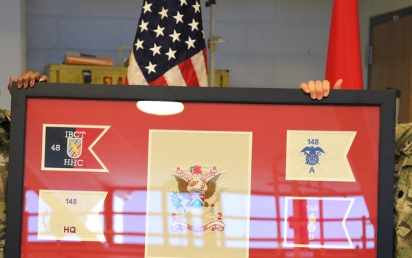Two U.S. Army soldiers in OCP (Operational Camouflage Pattern) uniforms and black face masks are holding a large framed shadow box display with a red background. The shadow box contains multiple military guidon pennants, unit patches, medallions, and a central coat of arms, appearing to be a retirement or farewell gift. A nameplate on the lower left reads 'CSM TAMARA E. MITCHELL,' suggesting this is a ceremony honoring her service.