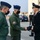 A formal military greeting ceremony is taking place outdoors, likely at a joint military installation in Italy. Two personnel in olive green flight suits and blue garrison caps are meeting with a senior officer in a dark formal Carabinieri uniform. All individuals are wearing face masks, suggesting this occurred during the COVID-19 pandemic period.