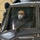 A bald man wearing glasses and a white KN95/FFP2 face mask is seated inside the cockpit of a military helicopter, likely during an inspection or tour. The helicopter appears to be parked inside a hangar, as indicated by the yellow-painted walls visible in the background. The aircraft shows riveted metal construction consistent with a military utility helicopter.