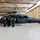 A military hangar contains a large dark-colored helicopter, likely an HH-60 Pave Hawk or similar Black Hawk variant, with a small group of civilians and uniformed personnel gathered near its open side door. The hangar is well-lit with a white and red structural framework visible overhead. A second helicopter is visible in the background on the left side of the hangar.