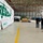 A group of four individuals, including military personnel in uniform and civilians, are gathered inside a large military hangar facility. The hangar features a prominent mural on the wall with the word 'RESCUE' in large green letters, accompanied by green footprints and a helicopter graphic. The space appears to be an active military aviation support facility with equipment and cargo visible in the background.