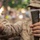 Two military personnel wearing desert camouflage uniforms and white face masks are holding objects in an outdoor, wooded setting. The foreground subject holds what appears to be a metal firearm component in one hand and a tape-wrapped cylindrical object in the other. The image is taken in shallow depth of field, keeping the hands and objects in sharp focus while the background is blurred.