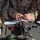 A military personnel wearing desert MARPAT (Marine Pattern) camouflage uniform is writing on a form or scorecard at what appears to be a shooting range or weapons qualification station. A rifle is positioned in the foreground on the surface, along with what appears to be a cleaning/lubricant bottle and other range materials. The scene suggests a weapons qualification or inspection documentation activity.
