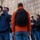 A group of people, likely tourists, are gathered in front of a large stone wall, possibly at a historic or religious site. One person in a blue jacket is actively photographing upward with a DSLR camera, while a central figure in an orange hoodie with a black backpack stands with their back to the camera. A man in a black jacket appears to be speaking or gesturing to the group, suggesting a guided tour setting.