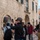 A group of tourists are gathered on a stone-paved street in what appears to be Dubrovnik's Old Town, Croatia, based on the distinctive limestone architecture. Several individuals are photographing the surroundings using smartphones and a DSLR camera. A shop called 'PLUS Souvenirs &amp; Fashion Jewelry' is visible in the background.