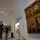 Museum visitors are viewing a large ornate gilded polyptych altarpiece featuring multiple religious figures painted in a Gothic or early Renaissance style. The setting appears to be a vaulted white-walled museum gallery, likely in a Mediterranean or Southern European institution. One visitor wearing a face mask is photographing the artwork while another consults a mobile device nearby.