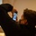A person wearing a black jacket and face mask is photographing what appears to be a religious icon or painting using a smartphone. The individual is angling the phone upward to capture the subject on the screen. The setting appears to be an indoor space, possibly a church, museum, or religious establishment.