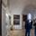 A group of museum visitors wearing face masks are viewing classical paintings in what appears to be a historic art museum with vaulted stone arches and white walls. One visitor is holding up a smartphone, likely photographing or recording the artwork. The space features multiple large oil paintings in gilded frames, a glass display case, and architectural elements suggesting a converted historic building.