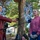 Two women are engaged in an outdoor interview, with one acting as a journalist holding a microphone labeled 'HRT' (Croatian Radio-Television) and a phone, while the other is being interviewed. The setting appears to be a public outdoor space with trees, buildings, and a colorful wall in the background. The interviewee is wearing a pink sweater and white gloves, suggesting a community or volunteer event context.