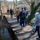 A group of people walking along a wooden boardwalk path through a natural area with bare winter trees and flowing water visible on both sides. The scene appears to be a nature park or tourist trail, likely during late autumn or winter based on the leafless trees and visitors' cold-weather clothing. The group consists of approximately 6-7 individuals moving away from the camera.