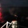 A nighttime naval scene showing a US Navy vessel with personnel operating what appears to be a mounted weapon system firing toward the water. Red flares or tracer rounds are visible illuminating the ocean surface in the distance. The image was taken from an elevated position on the vessel's deck.