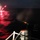 A nighttime maritime scene showing a US Navy vessel with personnel firing a flare gun or line-throwing device over open water. Red illumination flares are visible burning on the water's surface, casting a vivid red reflection across the waves. This appears to be a search and rescue exercise or distress signaling drill conducted at sea.