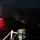 A nighttime maritime scene showing a US Navy vessel firing what appears to be flares or signal rounds over open water. Red illumination from flares reflects dramatically off the ocean surface to the left. At least one crew member is visible on the vessel's deck near what appears to be a weapon or flare launcher.