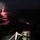 A nighttime maritime scene showing a US Navy vessel firing what appears to be a flare or signal rocket over open water. A bright red flare is visible in the distance reflecting off the ocean surface, while a second launch or firing event is captured mid-frame near the vessel's deck. The image is taken from an elevated position aboard the ship, showing deck railings and 'US NAVY' markings.