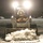 A person wearing a white face mask, fur-trimmed parka hood, and brown work overalls is operating a Bobcat skid-steer loader at night. The machine's headlights illuminate the scene as the operator pushes a pile of snow and ice with the front bucket attachment. The setting appears to be a parking lot or paved surface during or after a winter snowstorm.