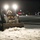 A person operating a compact track loader (likely a Bobcat) is clearing snow from a parking lot or open paved area at night. The operator is wearing a fur-trimmed parka hood and a face mask, suggesting cold weather conditions. The machine's headlights illuminate the snow-covered ground, with city lights visible in the dark background.