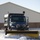 A dark blue Freightliner medium-duty dump truck equipped with a yellow front-mounted snow plow is actively clearing snow from a paved surface. Two occupants wearing face masks are visible in the cab. The truck also has a rear dump bed, likely loaded with sand or salt for road treatment.