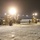 Two yellow motor graders are operating at night on a snow-covered surface, likely an airport tarmac or runway. The machines are actively clearing snow, illuminated by their own headlights and distant pole-mounted lights. A large aircraft is faintly visible in the background on the left side.