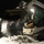 A compact track loader (likely a Bobcat) is being operated at night to clear snow from a paved surface. The machine's bucket is raised and actively dumping or pushing snow, with snow particles visibly spraying in the illuminated foreground. A single operator is visible inside the enclosed cab, working under artificial lighting.