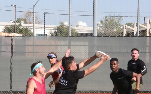A group of people are playing an outdoor flag football or ultimate frisbee game on a green turf field. A player in a black shirt is leaping to catch or deflect a white disc/ball while others in red and black jerseys compete around them. The game appears to be a casual or recreational co-ed match taking place on a fenced athletic field.