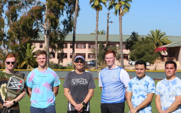 A group of eight people pose together on a green athletic field, likely at a military or sports facility based on the building and surroundings. One person is holding a WWE-style championship belt, and another holds what appears to be a toy sword. The group appears to be in athletic/casual wear, suggesting a recreational or team-building event.
