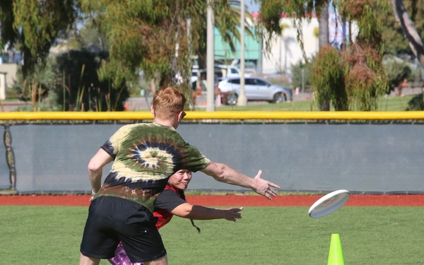 Two players are engaged in an ultimate frisbee game on an artificial turf field, with one player releasing or catching a white frisbee while the other defends closely. A green traffic cone marks a boundary or end zone on the field. The setting appears to be a recreational sports complex with a baseball outfield wall visible in the background marked '316'.