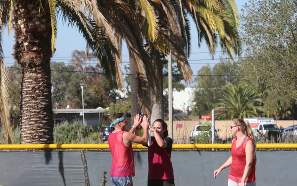Three athletes are on an artificial turf field, with two of them exchanging a high-five while a third stands nearby. They are wearing red/pink athletic gear and are surrounded by bright green training cones. The setting appears to be an outdoor sports facility bordered by a yellow outfield wall, with palm trees visible in the background.