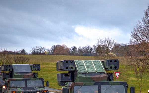 A military convoy of camouflage-painted HMMWV (Humvee) vehicles equipped with what appear to be Avenger air defense missile launcher systems is traveling along a paved road. The convoy consists of at least four vehicles moving in a single-file formation. The setting appears to be a rural European road, suggested by road signage including a yield/give-way triangle sign and road markers consistent with German or Central European highway standards.
