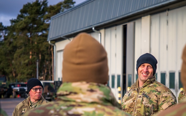 A group of military personnel in camouflage uniforms and cold-weather beanies are gathered outdoors near a large hangar or military building. A smiling soldier appears to be addressing or briefing the group, with another soldier standing nearby. The setting appears to be a military installation, likely in a cold-weather European environment based on the architecture and vegetation.