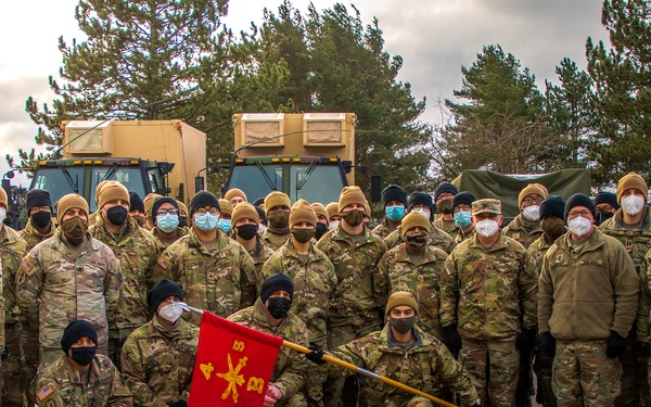 A large group of U.S. Army soldiers in camouflage uniforms and face masks pose for a unit group photo outdoors in cold weather conditions. Two soldiers in the front row kneel while holding a red unit guidon flag displaying what appears to be '4-3' with crossed cannons, indicating an artillery unit. Military vehicles including what appear to be HIMARS or similar tactical trucks are visible in the background.