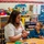 A female teacher or childcare worker is seated at a low table with a young toddler in a colorful early childhood classroom. The adult, whose shirt appears to read 'Fanani-Rodriguez,' is handling a small white object while the child watches attentively. The classroom is well-stocked with educational materials, toys, and supplies organized on shelving units.