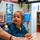 A young toddler wearing a blue hoodie with rainbow patterns sits at a small table in what appears to be a preschool or daycare classroom. The child is looking up attentively at an adult (partially visible on the left) who appears to be interacting with them. The classroom background shows colorful educational materials, storage shelves, bins, and learning toys.