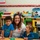 A smiling adult caregiver wearing a 'Tiger Cubs Child Care' shirt poses with two young children at a table in a colorful, well-equipped childcare classroom. The room is filled with educational toys, activity boxes, and learning materials organized on wooden shelving units. A decorative border and a 'write' label sign are visible on the wall behind them.